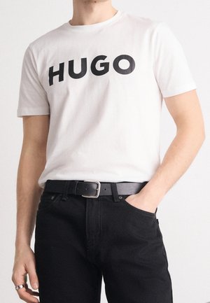 Person iført en hvid T-shirt med korte ærmer med "HUGO" i sorte bogstaver, sort bælte og sorte bukser, stående med den ene hånd i lommen.