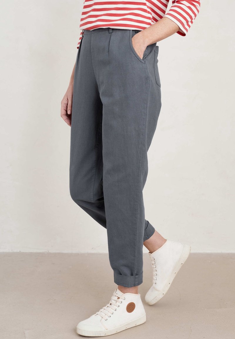 Personne portant un pantalon gris retroussé, une chemise rayée rouge et blanche, et des baskets blanches montantes, debout sur un sol beige.