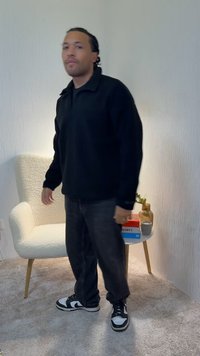 Crni pulover od runa, udoban kroj, uparen s tamnim trapericama i crno-bijelim tenisicama. Pozadina uključuje teksturirani zid i dekoracije.