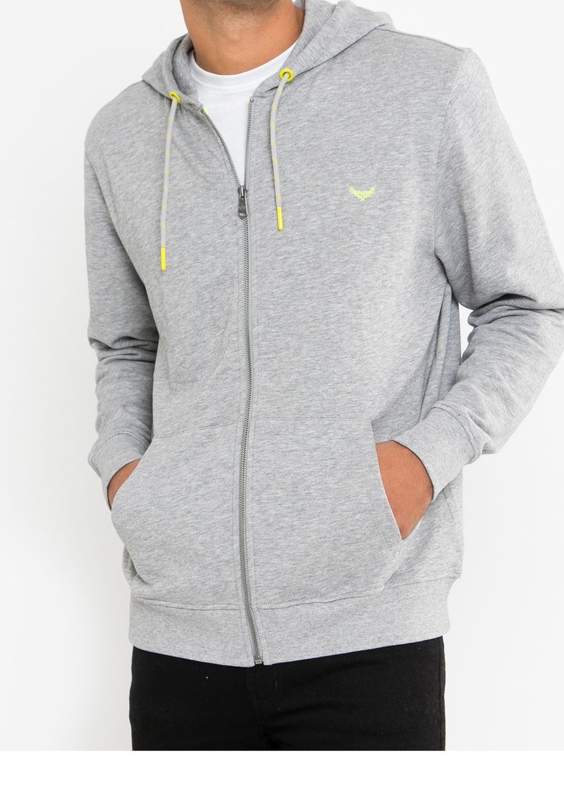 Sudadera con cremallera en color gris, confeccionada en material suave. Presenta una capucha con cordones y puntas amarillas, cierre de cremallera y bolsillos frontales. Diseño ligero.