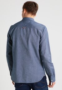 Mörkblå denimskjorta, långärmad, knappmanchett, detaljer med söm vid ryggen, liten logotyp nära kragen, slät textur.