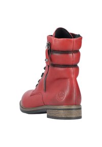 Botte de cheville en cuir rouge avec un col rembourré, des lacets noirs, une fermeture éclair sur le côté et un petit talon en bois. Texture lisse avec des détails de coutures subtiles.