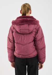 Bordeaux pufferjack met een dikke, pluche kraag en chevronstiksel. Voorzien van een capuchon en elastische manchetten, gecombineerd met zwarte broek.