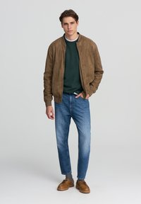 Giubbotto bomber in camoscio tan sopra un maglione verde scuro, abbinato a jeans blu chiaro e scarpe marroni. Caratteristiche includono polsini e orlo a coste.