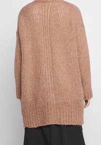 Person, die einen lockeren, strukturierten, hellbraunen Strickpullover trägt, von hinten über einem schwarzen Rock oder einer Hose betrachtet.