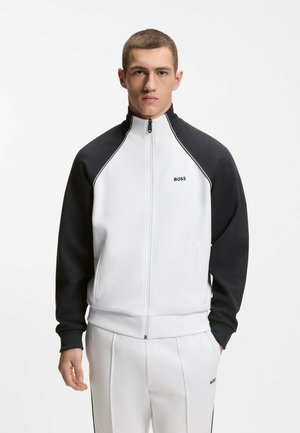 Joven con chaqueta blanca y negra con cremallera y pantalones blancos con el logo de BOSS, de pie con las manos en los bolsillos contra un fondo liso.