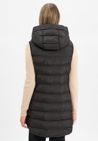 Schwarze Steppweste mit horizontaler Steppung, Kapuze und mittellangem Schnitt, getragen über einem cremefarbenen Pullover, mit glatter, glänzender Textur.