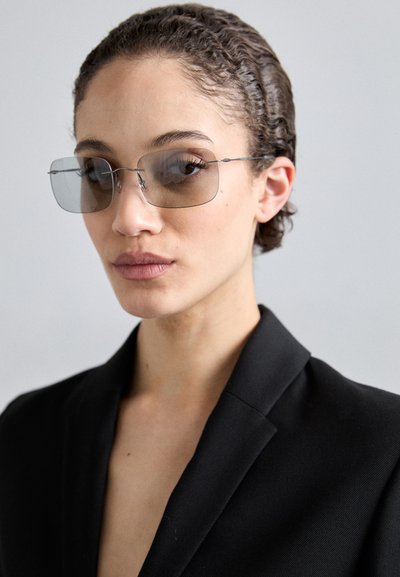 Lunettes de soleil avec des verres rectangulaires surdimensionnés de couleur gris foncé. Monture fine en métal avec des détails minimalistes. Portées avec un blazer noir.