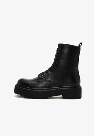 Botín de cuero negro hasta el tobillo con superficie texturizada, punta redonda, ojales de metal y suela de goma gruesa. Presenta una tira en la parte trasera.