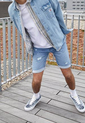 Person lehnt sich nach hinten, trägt eine hellblaue Jeansjacke, ein weißes T-Shirt, zerrissene Jeansshorts, weiße Socken und gemusterte Sneakers auf einem Holzdeck.