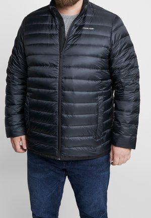 Mand iført en sort Calvin Klein quiltet dunjakke over en grå sweater og blå jeans, stående op ad en ensfarvet baggrund.