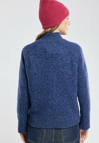 Un pull tricoté bleu avec un col montant, un ourlet côtelé et un tissu texturé. Un bonnet tricoté rose est porté sur la tête.