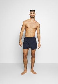 Calções de banho azul-marinho feitos de um tecido leve, com um cós elástico e um corte descontraído, apresentados por um modelo masculino sem camisa.