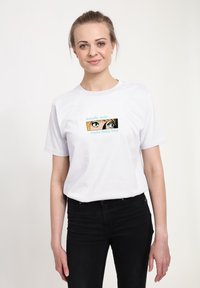 Hvid bomulds T-shirt med korte ærmer, der har et trykt grafisk design af øjne og tekst i grøn og blå. Bæres med sorte jeans.