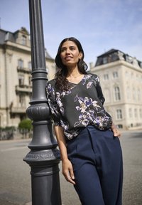 Blusa nera a fantasia floreale con maniche a sbuffo abbinata a pantaloni blu navy a vita alta, in piedi accanto a un lampione in un ambiente urbano.