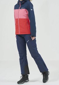 Giacca da sci isolata con pannelli rosa, rossi e blu navy, dotata di cappuccio e tasche con zip. Abbinata a pantaloni da sci blu navy e stivali neri.