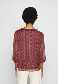 Blusa rossa con motivo, caratterizzata da un collo arricciato, maniche a tre quarti e un tessuto leggero e trasparente con righe verticali.