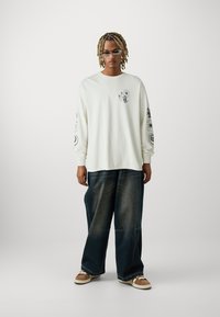 Tricou alb cu mânecă lungă, decorat cu desene grafice negre, asortat cu blugi negri largi și adidași maro cu tălpi albe.