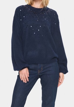 Marinblå stickad tröja med avslappnad passform, prydd med paljetter över bröstområdet; matchad med mörkblå jeans.