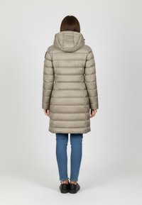 Beige puffatakki, jossa on huppu, tikattu pinta, polvipituinen malli, näkyy takaa. Yhdistetty sinisiin farkkuihin ja mustiin kenkiin.
