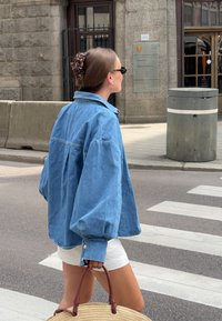 Hellblaues, übergroßes Jeansjackett mit Puffärmeln, weiße Shorts und eine geflochtene Strohtasche, während er auf einer Stadtstraße spaziert.