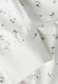 Tissu blanc à motif floral avec des fleurs rose pâle et des feuilles vertes. Le poignet présente une texture lisse et une finition côtelée.
