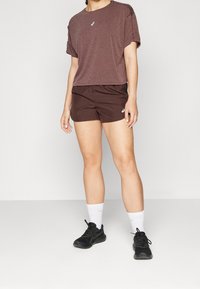 Camiseta atlética de manga corta color burdeos con detalles en los hombros, combinada con pantalones cortos marrones. Zapatillas deportivas negras y calcetines blancos completan el look.