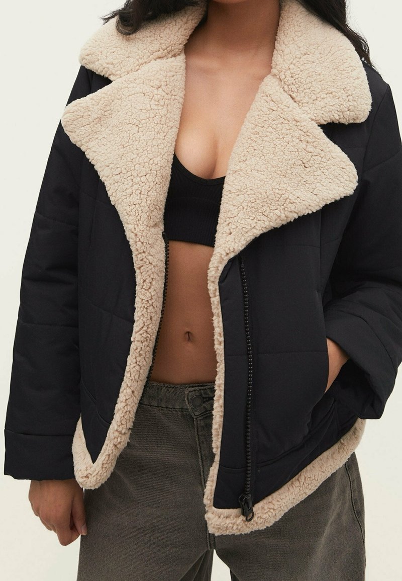 Femme portant une veste courte noire doublée de fourrure beige, dézippée pour montrer un haut court noir et un pantalon vert olive.