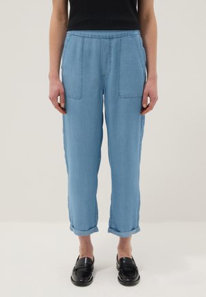 Persoon die lichtblauwe omslagbroek met voorzakken en zwarte loafers draagt, staand tegen een eenvoudige achtergrond.