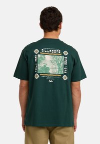 Camiseta de algodón verde oscuro con un estampado gráfico en la parte posterior que presenta diseños geométricos en crema y amarillo, y una imagen central de árboles.
