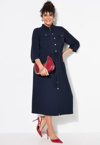 Marineblaues Hemdblusenkleid mit Dreiviertelärmel und Taillengürtel. Verfügt über zwei Brusttaschen und goldene Akzente. Kombiniert mit einer roten Handtasche und hohen Schuhen.