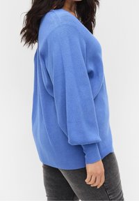 Hellblauer Strickpullover mit V-Ausschnitt, strukturiertem Stoff und übergroßen Ärmeln. Kombiniert mit dunkelgrauen Jeans, die eine entspannte Passform zeigen.