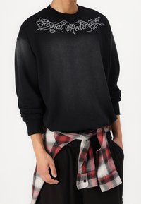 Persona che indossa una felpa nera con la scritta "Eternal Redemption", pantaloni neri e una camicia a quadri rossi e bianchi legata in vita, su sfondo bianco.