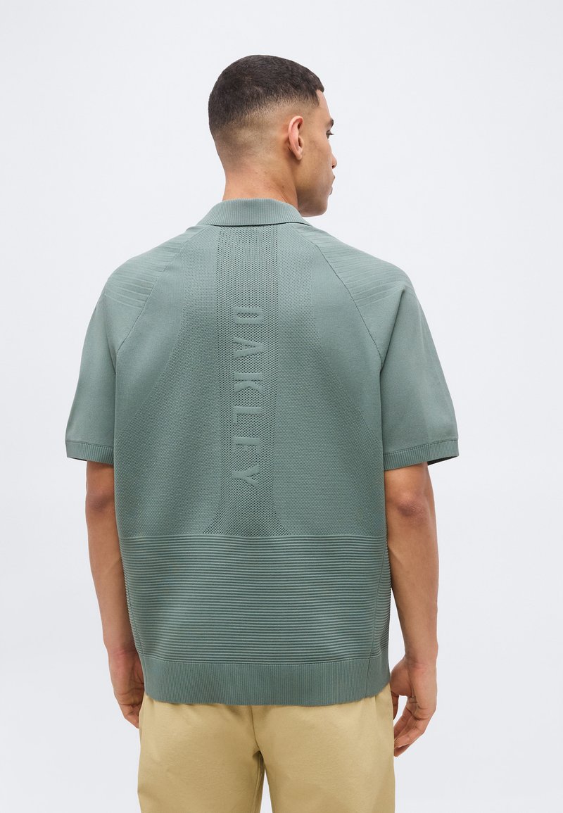 Άνδρας που στρέφεται προς τα πίσω φοράει ένα αμυδρά πράσινο κοντομάνικο πλεκτό πουκάμισο με την επιγραφή "OAKLEY" ανάγλυφη κάθετα στην πλάτη, συνδυασμένο με μπεζ παντελόνι.