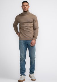 Brauner Rollkragenpullover aus Strickmaterial, kombiniert mit hellblauen Jeans und weißen Sneakern mit beigen Sohlen.
