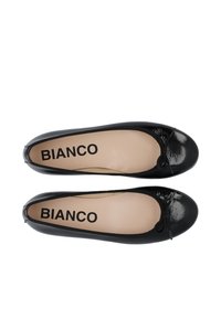 Pantofole di vernice nera con punta tonda, un piccolo dettaglio a forma di fiocco e il nome del marchio "BIANCO" stampato su una soletta color nude.