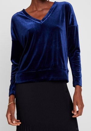 Kobieta ubrana w niebieską aksamitną bluzkę z długim rękawem i dekoltem w serek oraz czarną plisowaną spódnicę z bocznymi guzikami na jednolitym tle.