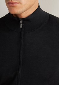 Svart zip-up tröja med hög krage, med en slät stickad textur och liten dragkedjedetalj vid halsen. Inga synliga mönster eller accenter.