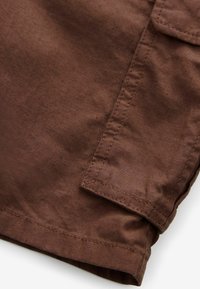 Brauner Stoffabschnitt eines Kleidungsstücks mit einer genähten Klappentasche und sichtbaren Nahtdetails.