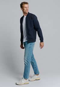 Marine bomberjack met gestructureerde stof, ritssluiting aan de voorkant, zijzakken en geribde manchetten. Gecombineerd met lichtblauwe jeans en witte sneakers.
