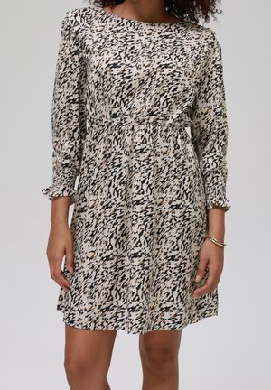 Kobieta ubrana w białą sukienkę z długim rękawem do kolan z abstrakcyjnym wzorem w czarne i brązowe plamy oraz złotą bransoletką na lewej ręce.