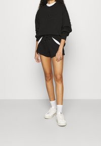 Černá uvolněná mikina se zvýrazněným logem, spojená s černými šortkami na šňůrku. Bílé tenisky s texturovanou podrážkou a kotníkové ponožky.