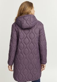 Quiltad lila lång kappa med huva, med ett mönster av vågiga linjer och en mjuk textur. Synliga sömmar och en avslappnad passform.
