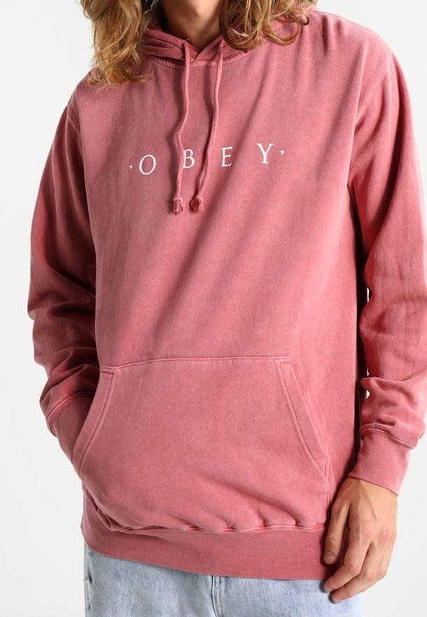 Personne portant un sweat à capuche rose pâle délavé avec poche avant et texte blanc "OBEY", associé à un jean bleu clair.