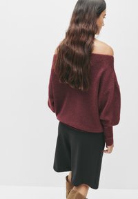 Bordeaux off-the-shoulder trui met een textuurgebreide stof, oversized pasvorm, geribbelde manchetten, gecombineerd met een zwarte knielange rok en beige laarzen.