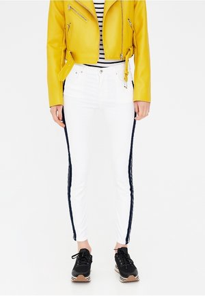 Personne portant un pantalon blanc avec des bandes latérales noires, une veste jaune zippée par-dessus un haut à rayures noires et blanches, et des baskets noires.