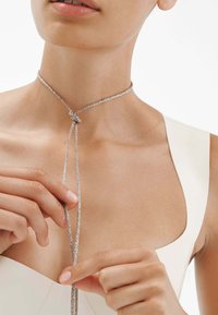 Collar plateado hecho de finos hilos, con un diseño de nudos y dos hilos colgantes. Textura metálica, estilo minimalista.