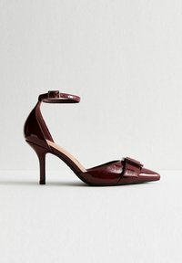 Scarpa con tacco alto in pelle verniciata bordeaux, con punta affilata, cinturino alla caviglia con fibbia e fibbia quadrata decorativa nella zona delle dita.