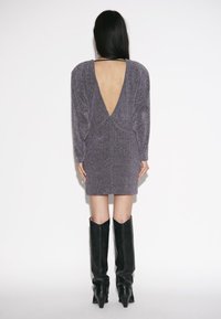 Vrouw met lang donker haar draagt een fonkelende paarse minijurk met diepe V-rug en zwarte kniehoogte laarzen met hakken, staand tegen een witte achtergrond.