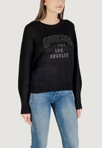 Schwarzer Pullover mit "GUESS" und "LOS ANGELES" aus metallischen Strasssteinen, entspannte Passform, gerippte Bündchen und Saum, getragen mit blauen Jeans.
